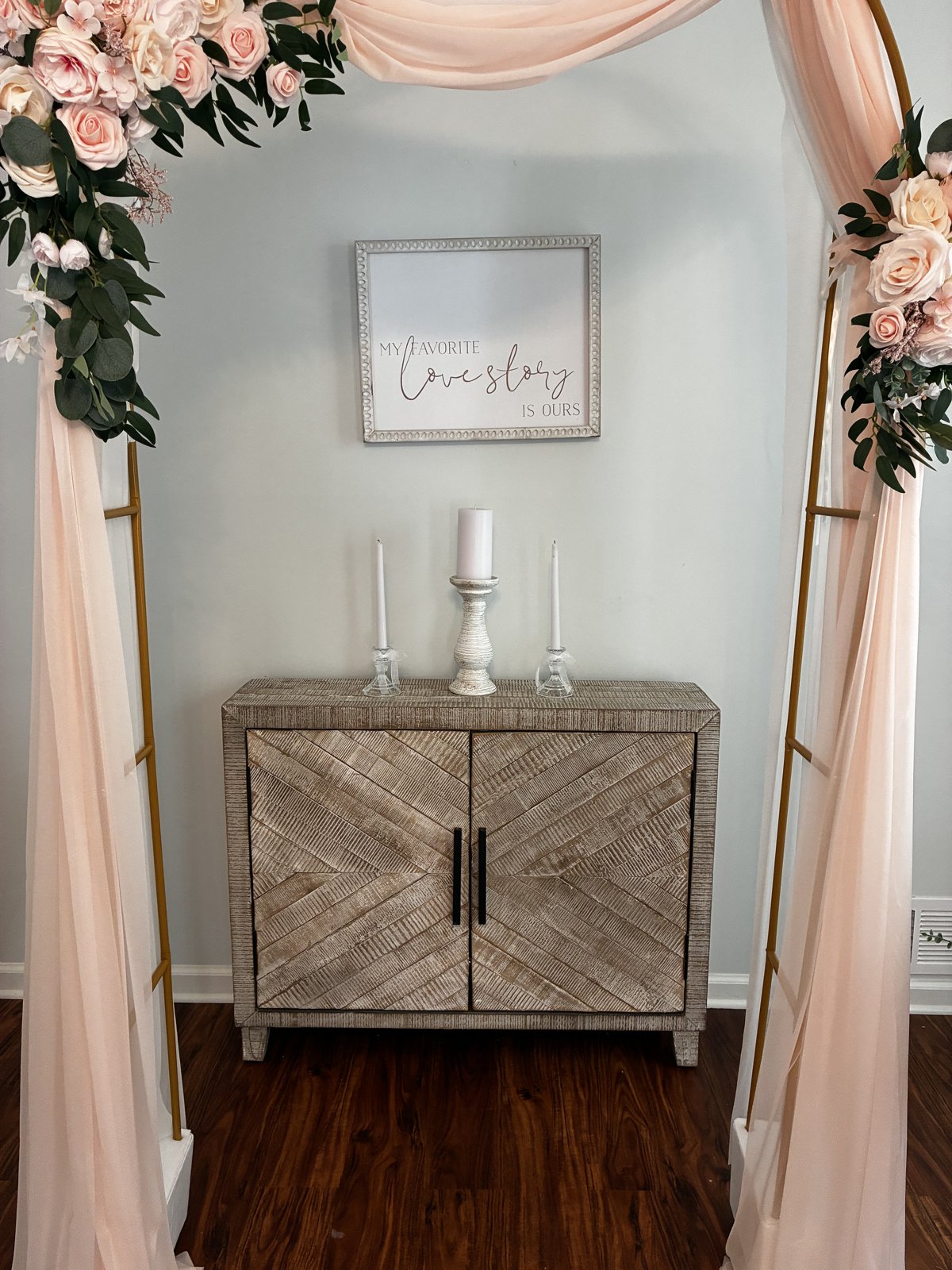Unity candles on a wooden cabinet beneath a My Favorite Love Story Is Ours sign, framed by the blush draped arch